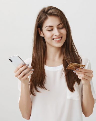 Pleased girlfriend sending money via internet banking. Indoor portrait of satisfied charming woman holding smartphone and credit card, typing account number while making purchase online. Copy space